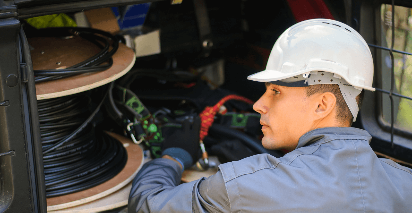 A young man wearing a white hard hat looks over his electricians equipment and a reel of black cable in the back of his truck.