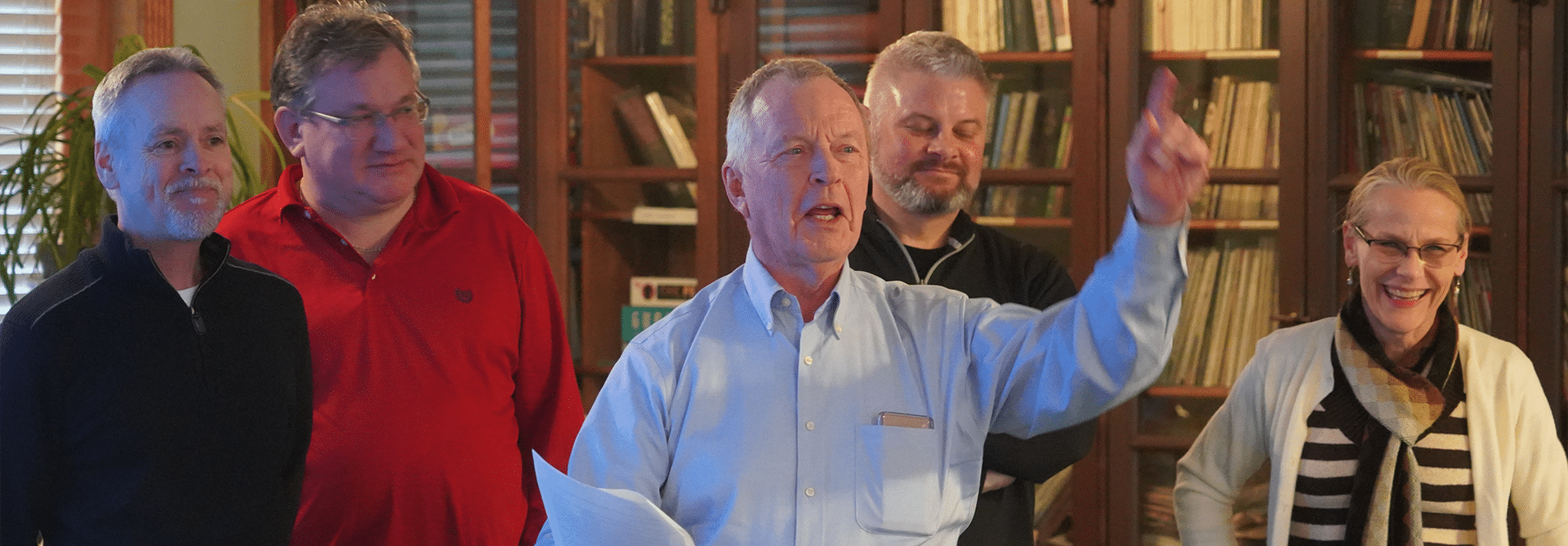 A man standing in front of his group during a group presentation, gestures dramatically by pointing to the sky. The other group members look amused.