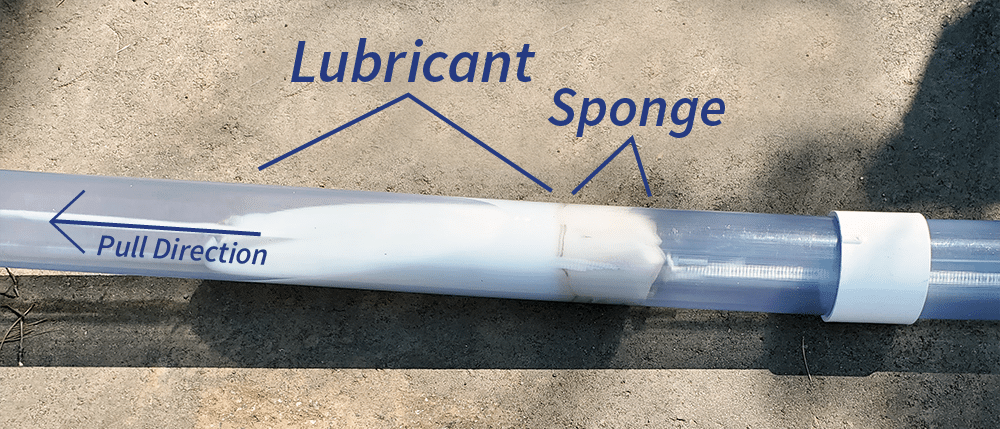 A clear plastic conduit lays on a concrete slab. Inside the conduit is a pile of white liquid lubricant with a sponge attached with a rope on both ends. When pulled through the conduit, the sponge spreads the lubricant evenly around the conduit. Text on the image labels the "lubricant", "sponge", and an arrow with the words "Pull direction", show the direction the sponge is pulled.