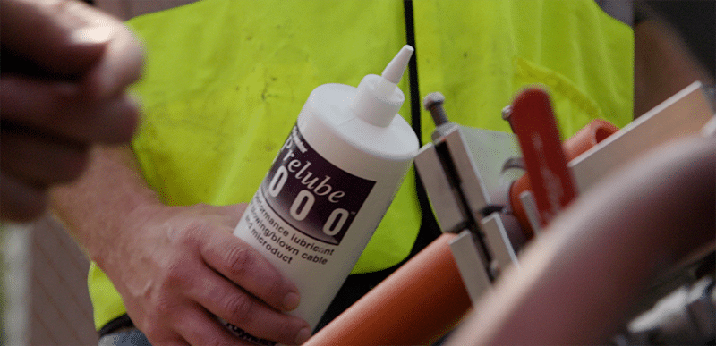 Un trabajador con un chaleco de seguridad de neón sostiene una botella de Polywater Prelube para su uso en el cable de fibra óptica que se sopla en un ducto naranja.