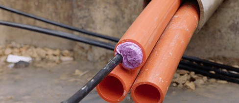 Trois conduits orange sortent d’un plus grand conduit dans un mur en béton. L’un des conduits est scellé avec une mousse violette - Polywater AFT - et un câble à fibre optique noir en sort.