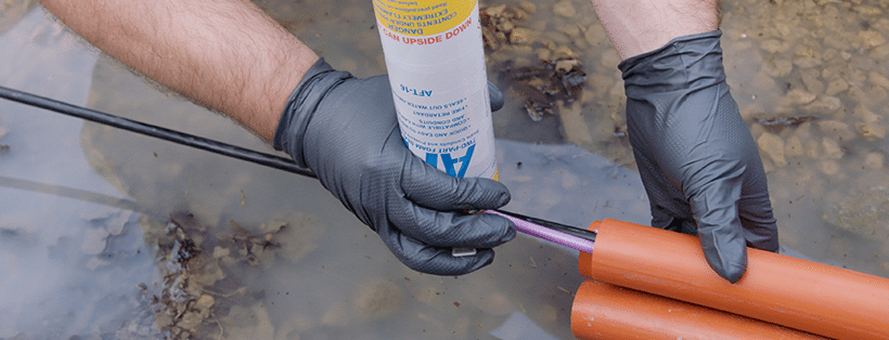 black-gloved hand holding an aerosol can of Polywater AFT foam sealant, deploys the sealant into an orange conduit used for fiber optic cable