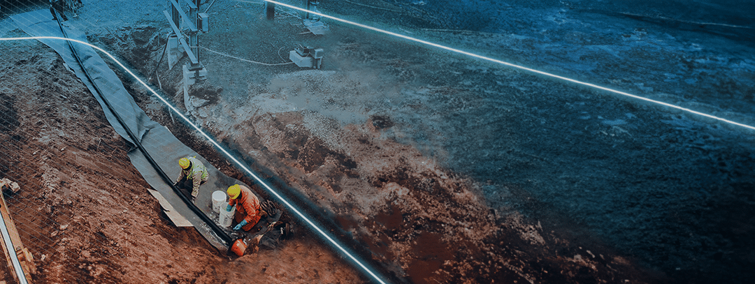 Two men lubricate an electrical cable in a trench as it goes into a conduit underground. A few workers help the cable off the reel in the background.