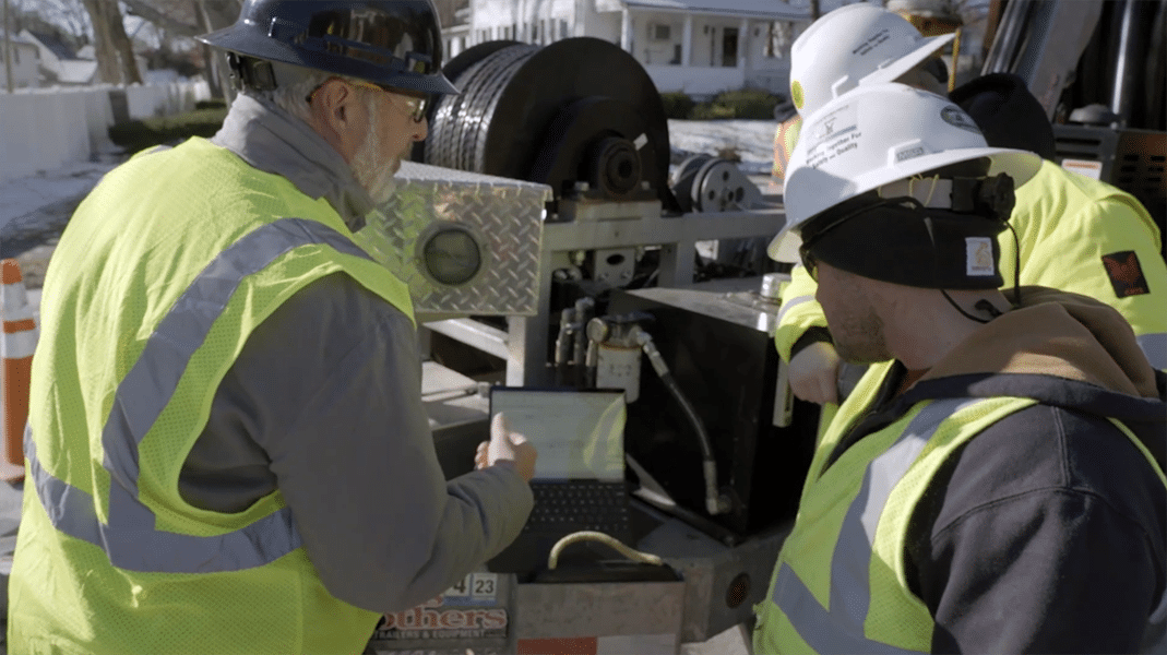 Workers in hard hats and green safety vests look at a lap top displaying the Polywater Pull-Planner on the back of their work truck.