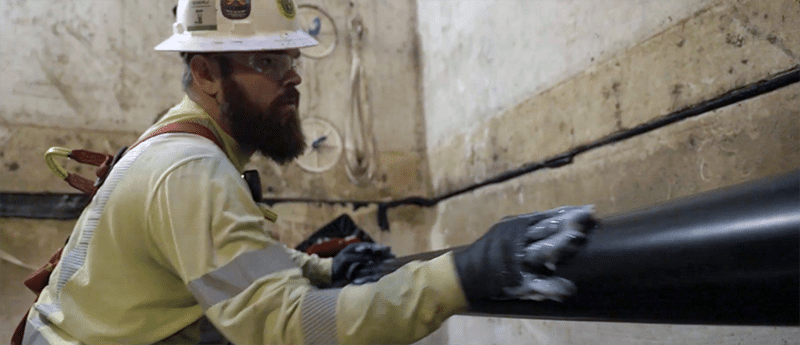 Man in an underground vault, hand-lubricates a large cable as its pulled through the conduit.